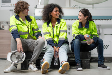 Break time. Group of diverse engineer people sitting together take a break and talking. Young multiracial woman and colleagues technician relax after job in factory. Manufacturing employee lifestyle. - Powered by Adobe