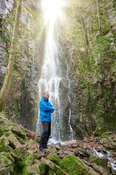 Tourist attraction of Germany - falls of Burgbach Waterfall near Schapbach, Black Forest, Baden-Wurttemberg, Germany. Man hiker in blue jacket standing on stone and looks at flow of falling water