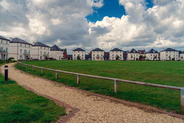 Fototapeta na wymiar Authentic street in the English countryside, green field and gravel path in the foreground, English country houses and cloudy sky, summer day. Sustainable architecture and sustainable design