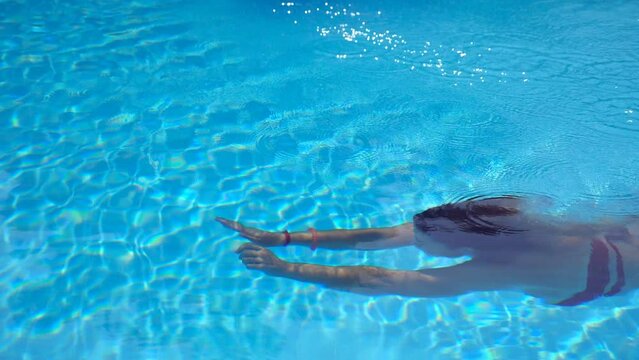 Unrecognizable woman swimming under water in pool with transparent clear water. Young girl relaxing during summer vacation swimming in basin of hotel. Concept of summer vacation. Slow motion Side view