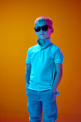 Portrait of little boy, child in casual clothes and sunglasses standing against yellow studio background in neon light. Concept of childhood, lifestyle, emotions, education, fashion, care, ad