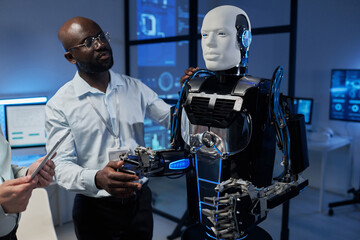 Engineer controlling robot from digital tablet with his colleague holding his robotic arm and...