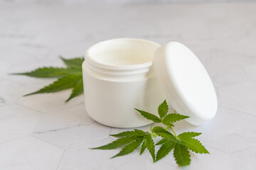 Opened white cream jar with a lid near cannabis leaves close up. Cosmetic Mockup