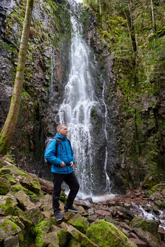 Tourist attraction of Germany - falls of Burgbach Waterfall near Schapbach, Black Forest, Baden-Wurttemberg, Germany. Man hiker in blue jacket standing on stone and looks at flow of falling water