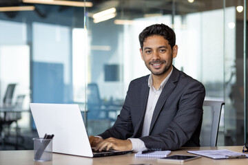 Portrait of young arab businessman, man smiling and looking at camera while sitting inside office, boss in business suit at workplace using laptop. - obrazy, fototapety, plakaty
