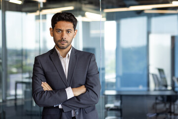 Portrait of mature serious businessman, arab with crossed arms thinking looking at camera, boss in business suit inside office at workplace standing near window.