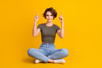 Full length photo of sweet shiny girl dressed khaki top sitting legs crossed pointing two fingers up empty space isolated yellow color background