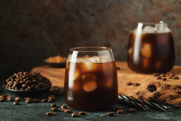 Ice coffee in a tall glass with cream poured over, ice cubes and beans on a dark concrete table.