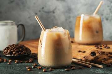 Ice coffee in a tall glass with cream poured over, ice cubes and beans on a dark concrete table.