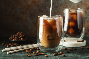 Ice coffee in a tall glass with cream poured over, ice cubes and beans on a dark concrete table.