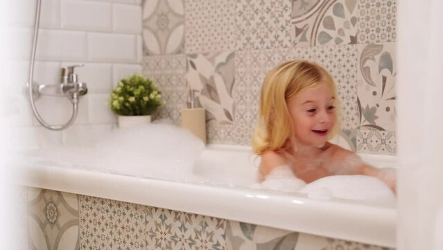 Cute funny caucasian blonde three years old girl taking bath with white foam bubbles, playing,smiling , having fun.Carefree childhood, kid body care concept. 