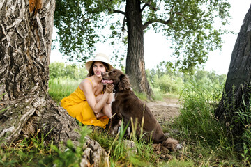 Pretty young woman in yellow dress and a hat sitting under the tree with dog. Female relax in hot summer day.