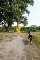 Pretty young woman in yellow dress and a hat sitting under the tree with dog. Female relax in hot summer day.