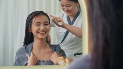 Medium shot of hairdresser combing and blowing dry hair of young female client in front of mirror at Beauty Salon