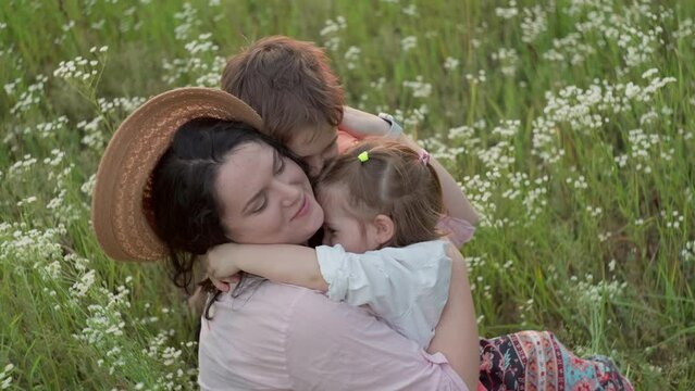 Son and daughter hug their mother while sitting in the grass. Happy moments of life, lifestyle of motherhood and children's love, smiles. High quality 4k footage