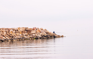 Front view, very, far, distance of, a lone fisherman with long fishing pole, sitting on rock jetty,...