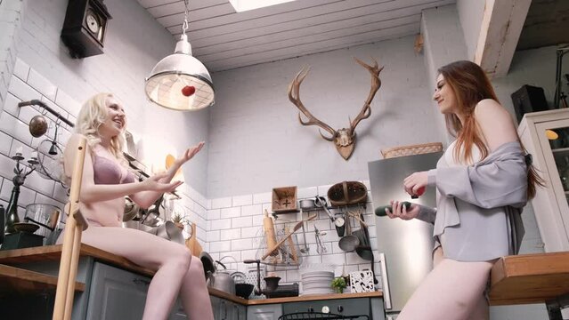 Two young, beautiful girls in underwear and shirts are having fun in the kitchen. They throw vegetables at each other. Healthy food concept
