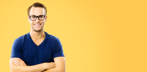 Portrait image of handsome happy man wearing in glasses, casual smart blue clothing, with crossed arms. Orange yellow colour background, with copy space. Male model in eyewears at studio.