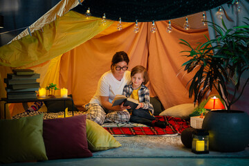 A higher close up camera shot of a mother reading out a storybook to her son that is sitting next to her in his comforting blanket fort