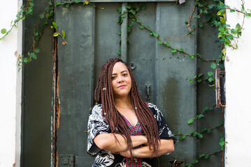 Portrait of a pretty woman with braids in her hair with her arms crossed against a green door with green plants.