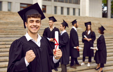 Young smiling joyful guy student in university graduate gown and diploma in hands. Happy man making yes gesture outdoor on the background of classmates. Graduation and education concept.
