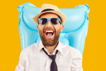 Cheerful handsome bearded business man in office shirt, tie, sunhat and sunglasses with inflatable beach water mattress laughs happily and has fun on summer holiday trip. Isolated on yellow background