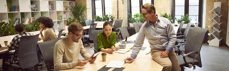 Sharing fresh ideas. Three young colleagues sitting at the desk in the modern coworking space, chatting and discussing new project, business people working together