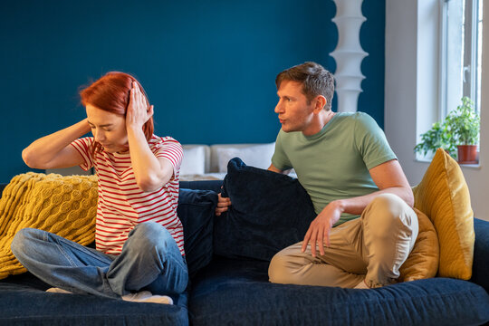 Unhappy couple quarrelling fighting at home sitting on couch. Annoyed husband shouting at upset woman closing ears. Woman ignore emotional pushy husband. Difficult family conflict, divorce break up. 
