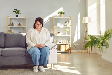 Stressed overweight woman sitting on sofa in living room at home. Troubled plus size woman looking...