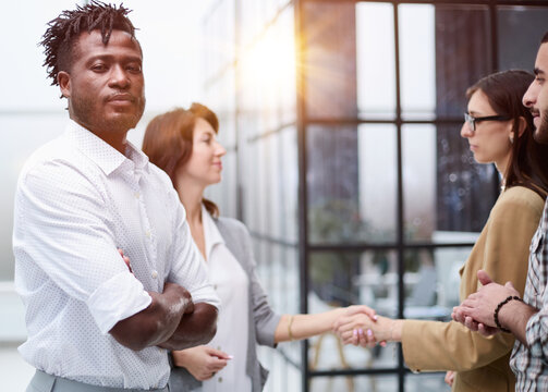 a serious black guy stands with his arms crossed against the background of colleagues shaking hands