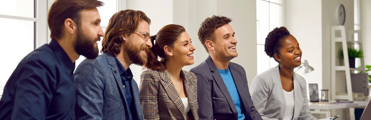Multicultural happy peoples men and women sits in row watching presentation or speech by colleague at corporate team building event dressed in business suits, located in room. Panoramic portrait
