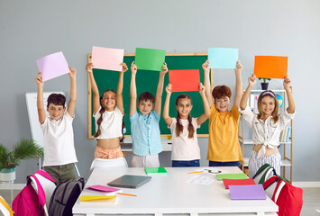 Cheerful school children standing in row in classroom and holding different colorful mockup...