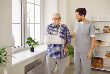 Young doctor or nurse in uniform supporting an injured senior patient. Friendly caregiver talking...