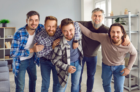 Portrait of a male bearded friends looking cheerful at the camera and smiling having fun at home. Full length photo of group of brutal friendly men in casual clothes hugging indoors on the party.