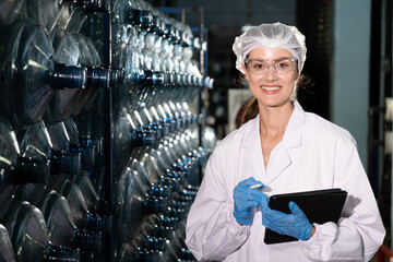 Caucasian women scientist with lab cote inspect quality of drinking water inside of the production line factory while using digital tablet to check cleanliness and standard, quality assurance