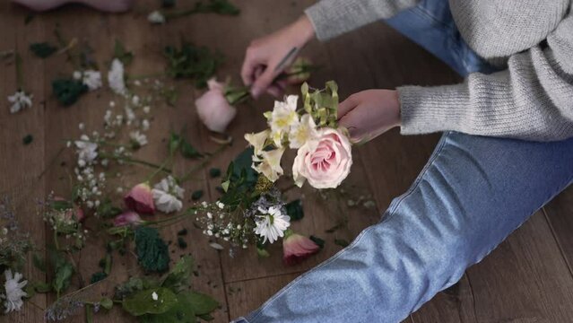 Live camera moves up as unrecognizable frustrated woman gathering broken bouquet in slow motion. Young Caucasian stressed depressed lady sitting on floor with flowers after fight