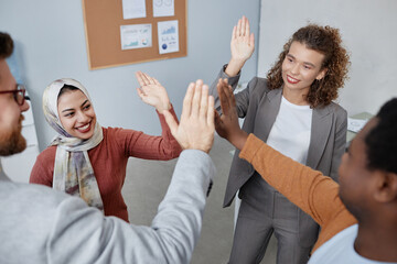 Happy young intercultural colleagues in smart casualwear giving each other high five while standing in circle in front of camera in office - obrazy, fototapety, plakaty