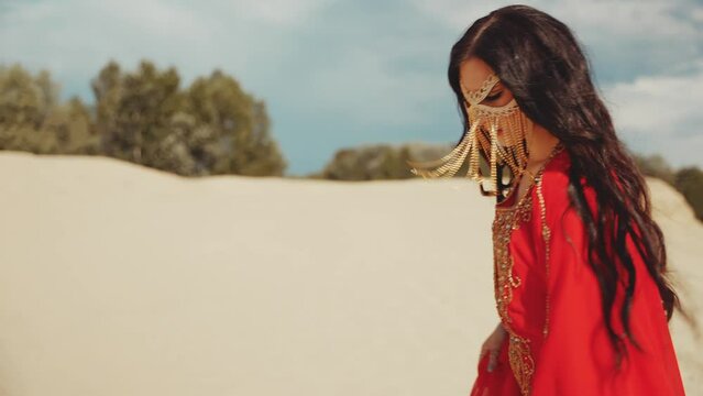 Arabian oriental woman walks in desert beauty face hidden by facekini Niqab golden mask veil chain. red gold dress long hair fly in wind. sexy girl arab style fashion model. blue sky nature Dubai UAE.