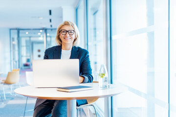 Portrait of 50's confident mature businesswoman looking at camera, middle-aged experienced senior female professional working on laptop in open space office. Female entrepreneur working remotely - obrazy, fototapety, plakaty