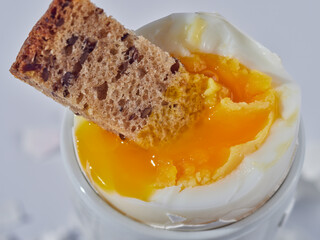 A piece of bread is dipped into an egg with liquid yolk in a white egg cup