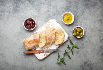 Sliced fresh ciabatta on cutting kitchen board, green and brown olives, olive oil with rosemary,...