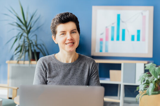 Portrait of smiling neutral gender middle aged woman at office desk with laptop and chart board with financial graphs on background. Business analysis concept. Data reporting. financial performance
