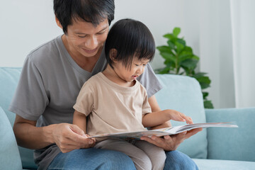Fototapeta premium Happy Asian father relax and read book with baby time together at home. parent sit on sofa with daughter and reading a story. learn development, childcare, laughing, education, storytelling, practice.