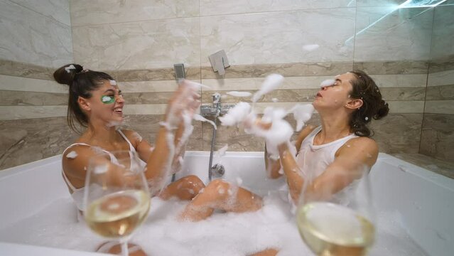 two pretty girls in white wet tops have fun in the bath with soap splashing water