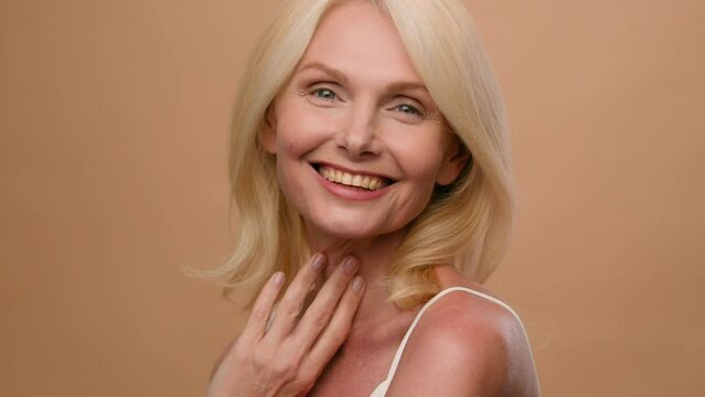 Portrait middle aged happy mature Caucasian woman senior 50s model flirty smiling at camera on beige studio background anti-aging beauty skin care procedures face lifting anti-wrinkle plastic surgery