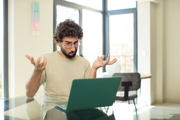 young adult bearded man with a laptop looking puzzled, confused and stressed, wondering between...
