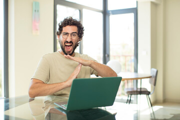young adult bearded man with a laptop looking serious, stern, angry and displeased, making time out sign