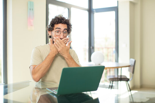 young adult bearded man with a laptop covering mouth with hands with a shocked, surprised expression, keeping a secret or saying oops