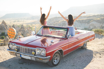 two beautiful girl in a retro dress at sunrise happy posing on the background of the mountain landscape in Cappadocia near a red retro car