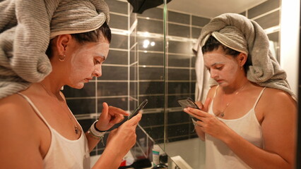 Woman night routine after bath covered with face mask checking phone in front of bathroom mirror